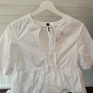 Vit blus med knyt från DIVIDED XS - Säljer en söt vit blus från hm i storlek XS. Blusen har knyt framtill vid halsen, korta ärmar och en volang nedtill. Tillverkad i mjuk bomull, perfekt för vår och sommar. Passformen är normal och toppen är enkel att matcha.