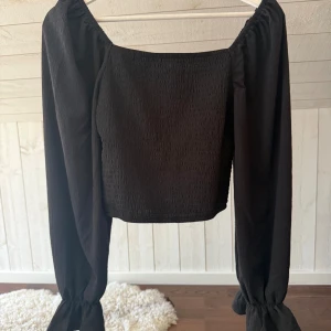 Svart croppad blus med puffärm - Säljer en svart croppad blus med fyrkantig urringning och långa puffärmar med resår vid ärmslut. Toppen har en smockad struktur på kroppen och är perfekt för dig som gillar trendiga och stilrena plagg. Passar snyggt till både jeans och kjol.