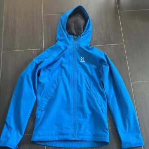 Haglöfs jacka - Snygg blå regnjacka från Haglöfs i storlek S. Jackan har huva, dragkedja framtill och två snedställda fickor. Perfekt för blåsiga och regniga dagar. Klassisk logga på bröstet och justerbar huva för extra skydd. Nypris 1800 kr. Skick 8/10