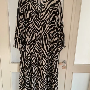 Zebramönstrad långklänning från Rainbow - Snygg långklänning från Rainbow med svartvitt zebramönster och v-ringad hals. Klänningen har volangdetaljer och trekvartsärmar, vilket ger en cool och avslappnad vibe. Tillverkad i mjuk polyester som känns skön mot huden.