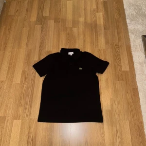 Svart pikétröja från Lacoste - Klassisk svart pikétröja från Lacoste med den ikoniska krokodilloggan på bröstet. Tröjan har krage, knäppning framtill och korta ärmar. Perfekt för en clean och stilren look. Tillverkad i mjuk bomull som andas.