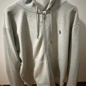 Grå hoodie från Polo Ralph Lauren - Säljer en stilren ljusgrå hoodie från Polo Ralph Lauren med dragkedja framtill, huva med snörning och klassisk broderad logga på bröstet. Tröjan har kängurufickor och är tillverkad i mjuk bomullsmix. Perfekt för chill dagar eller när du vill vara casual.