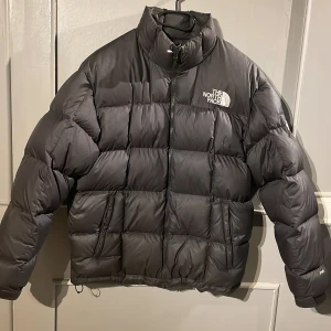 Svart pufferjacka från The North Face - Säljer en svart pufferjacka från The North Face med hög krage och synlig logga på bröstet och ärmen. Jackan har quiltad design, dragkedja framtill och två sidofickor. Perfekt för kalla dagar och har en riktigt schysst streetstil.