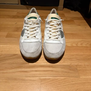 Vita och gröna sneakers från Philippe Model - Snygga vita sneakers från Philippe Model med gröna detaljer på hälen och grå mocka på tån och sidorna. Skorna har snörning, platt sula och en ikonisk sköld på sidan. Ovandelen är i textil och skinn, vilket ger en sportig och fräsch look.