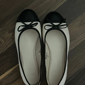 Svartvita ballerinaskor med rosett - Snygga ballerinaskor från Primark i svart och vitt med rund tå och dekorativ rosett framtill. Skorna har svart tåparti och kant, vilket ger en klassisk look. Perfekta att matcha med både jeans och kjol för en stilren vibe.