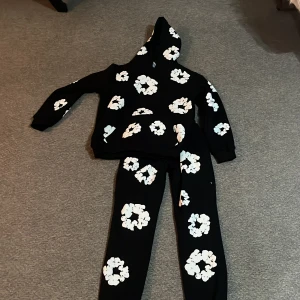 Svart hoodie med vita blommor Denim Tears - Svart hoodie från Denim Tears i storlek M med stora vita blommönster över hela tröjan. Tröjan har huva och en klassisk känguruficka framtill. Perfekt för dig som vill sticka ut med en unik och trendig streetwear-look. 900 för hela set men pris kan diskuteras 