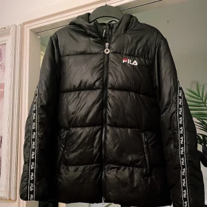 Svart pufferjacka från FILA med huva passar även som S - Svart pufferjacka från FILA med huva och dragkedja framtill. Jackan har logoband längs ärmarna och broderad FILA-logga på bröstet. Fylld för extra värme och har två fickor med dragkedja. Perfekt för kalla dagar och riktigt snygg streetstil.