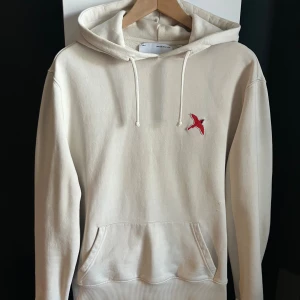 Beige hoodie från Axel Arigato - Säljer en stilren beige hoodie från Axel Arigato med broderad röd fågel på bröstet. Tröjan har huva med snörning och en stor magficka. Perfekt för dig som gillar minimalistisk design. Hoodien är i 9/10 skick, endast liten defekt över fickan. (Se bild 4) Storlek S