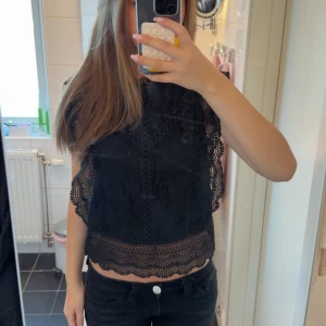 Svart spetsblus med volangdetaljer - Säljer en svart blus i spets med fina volangdetaljer längs ärmar och nederkant. Blusen är ärmlös och har en croppad passform. Baksidan är helt i spets med blommigt mönster som ger en snygg och trendig look. Tror den är från only men minns inte