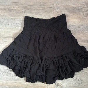 Svart volangkjol från Lager 157  - Svart kortkjol från 767 i storlek XS. Kjolen har en bred, smockad midja som ger en skön passform och volangdetaljer nedtill för extra flärd. Tillverkad i mjukt bomullsmaterial, perfekt för dig som gillar en feminin och chill stil.