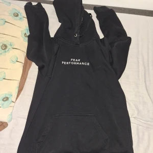 Svart hoodie från Peak Performance - Svart hoodie från Peak Performance med vit text på bröstet. Klassisk modell med huva, snörning och känguruficka. Tillverkad i mjuk bomulls- och polyestermix, perfekt för chill dagar eller när du vill ha en clean streetstyle.