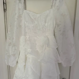 Vit kortklänning med volanger - Superfin vit kortklänning med puffiga långärmar och volanger i flera lager. Klänningen har ett subtilt blommigt mönster i tyget och en söt v-ringad urringning. Samt stretch i ryggen. Passar perfekt till studenten eller annan festlighet!!!!!Helt ny med prislapp kvar! Nypris 320 kr