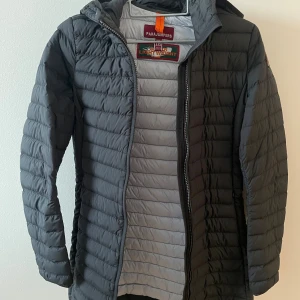 Svart dunjacka från Parajumpers - Säljer en svart, quiltad dunjacka från Parajumpers med huva och dragkedja framtill. Jackan har en smal passform och klassisk logga på ärmen. Perfekt för kyliga dagar och riktigt lätt att bära. Snygg och stilren design som funkar till det mesta.