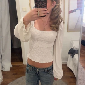 Vit smockad blus med puffärmar - Supersöt vit blus med smockad kropp och långa puffärmar. Toppen har en fyrkantig ringning och lätt volangkant. Passar till allt. Nyskick