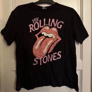 Mörk grå Rolling Stones t-shirt - Cool mörk grå t-shirt med klassiskt Rolling Stones-tryck framtill, med den ikoniska tungan i rosa och vitt. T-shirten har rund hals och korta ärmar passar även en m/l