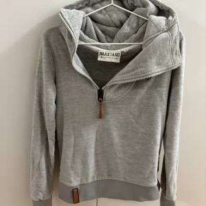 Grå hoodie från Naketano med half zip - Världens finaste och mysigaste gråa hoodie från Naketano med half zip och stor huva. Tröjan har långa ärmar, ribbade muddar och en mjuk insida. Perfekt för kyliga dagar och har snygga detaljer i läderimitation vid dragkedjan och nederkanten.