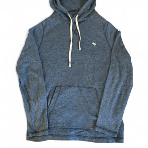 Blå hoodie från Abercrombie & Fitch | S - Mysig blå hoodie från Abercrombie & Fitch med vit dragsko och klassisk älglogga broderad på bröstet. Tröjan har känguruficka framtill, huva och är tillverkad i mjuk bomullsblandning. Perfekt för chill dagar eller när du vill ha en avslappnad stil.