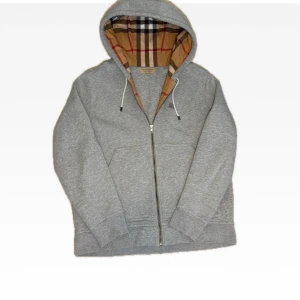Grå Burberry hoodie - Klassisk grå hoodie från Burberry med klassiskt rutigt foder i huvan. Tröjan har YKK dragkedja (stainless steel), snörning i huvan och broderad logga på bröstet. Tillverkad i mjuk bomull och har långa ärmar med ribbade muddar. Perfekt för en chill och stilren look. Nypris på dessa är ca7k så i denhär kvalitén är priset väldigt bra!