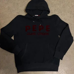 Svart hoodie från Pepe Jeans London - Svart hoodie från Pepe Jeans London med röd broderad logga på bröstet. Klassisk känguruficka framtill, huva med snörning och ribbade muddar. Regular fit och mjukt material som känns skönt mot huden. Perfekt för dig som gillar stilrena streetwear-plagg.