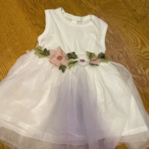 Vit kortklänning med blommor 9-12 mån/ 74 cm  - Superfin vit kortklänning med ärmlös design och fluffigt tyllkjol. Midjan är dekorerad med broderade blommor i ljusrosa och vitt samt gröna blad, vilket ger en söt och romantisk vibe. Perfekt för sommarens speciella tillfällen.