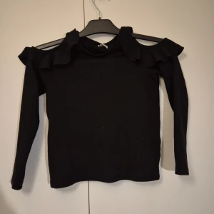 Svart långärmad topp med volang Lindex - Svart långärmad topp från Lindex med snygga volangdetaljer vid axlarna och öppna axlar för en cool look (den gör sig inte riktigt på galgen). Toppen är i mjuk bomull och har en slim passform.