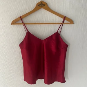 Vinröd linnetopp med smala axelband - Säljer en vinröd linnetopp med smala axelband och v-ringad hals. Toppen har en lätt glansig finish och är tillverkad i ett mjukt material som ger en lyxig känsla. Perfekt att styla med jeans eller kjol för en trendig look.