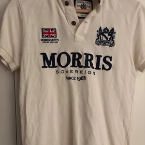 Vit Morris kortärmad piké - Vit piké från Morris med broderad logga, brittisk flagga och texten 'Morris Sovereign' framtill. Klassisk krage med knappar och korta ärmar. Tillverkad i bomull för en skön och stilren känsla.