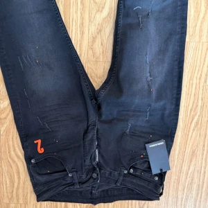 Svarta Dsquared2 - Snygga svarta jeans från Dsquared2 med orange '2' på fickan. Jeansen har distressed detaljer och raka ben. Klassisk femficksmodell med Dsquared2-logga vid gylfen. Perfekta för dig som gillar Dsquared2 jeans!