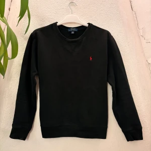 Svart sweatshirt från Polo Ralph Lauren - Svart sweatshirt från Polo Ralph Lauren med klassisk rund hals och broderad röd logga på bröstet. Tröjan har långa ärmar och ribbade muddar vid ärmslut och nederkant. Perfekt för en clean och stilren look.