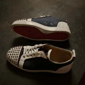 Christian Louboutin sneakers med nitar - Säljer ett par Christian Louboutin sneakers i vitt och mörkblått med ikoniska röda sulor. Skorna har denimliknande tyg, vita skinnpaneler och coola silverfärgade nitar på tån. Klassisk snörning och rund tå. Kommer med originalkartong och dustbags. 