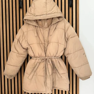 Beige pufferjacka med huva och snörning - Säljer en beige pufferjacka med stor huva och snörning i midjan för extra passform. Jackan har quiltad design och långa ärmar, perfekt för kalla dagar. 