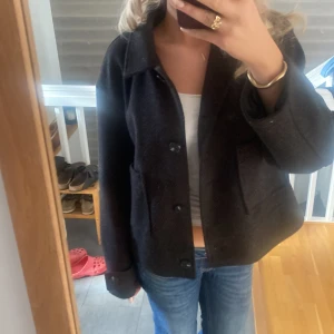 Kappa - Säljer en mörkbrun jacka i ull med klassisk krage och stora knappar framtill. Jackan har två rejäla fickor och en rak, något boxig passform. Perfekt för dig som gillar en stilren och enkel look.