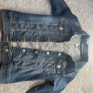 Jeansjacka från Lab Industries stl. XXS - Snygg blå jeansjacka från Lab Industries med klassiska knappar framtill och cool broderad logga på ryggen. Jackan har kontrastsömmar, bröstfickor med lock och knapp samt detaljer i metall. Perfekt för dig som gillar streetstyle och denim.