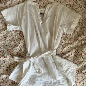 Vit blus med knyt  - Vit blus i bomull med korta ärmar, v-ringning och breda ribbade detaljer vid ärmslut och hals. Midjan markeras med ett knytband för en snygg siluett. Perfekt för en fräsch och stilren look.