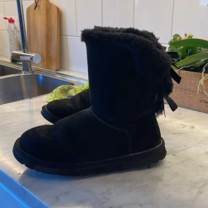 Svarta UGG boots med rosetter - svarta UGG boots i mocka med fluffig insida och snygga rosetter bak på skaftet. Klassisk rund tå och platt sula, perfekta för kalla dagar. Utsidan är i mjuk mocka och insidan är fodrad med fårskinn för extra värme.