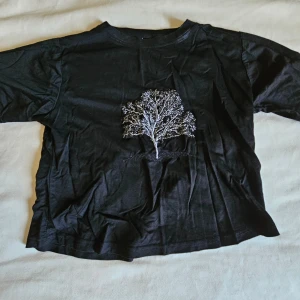 Svart t-shirt med broderat träd - Svart t-shirt med ett snyggt broderat träd i vitt på bröstet. Klassisk rund hals och korta ärmar. Perfekt för dig som gillar naturmotiv och vill ha en unik detalj på din outfit.