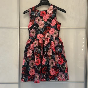 Blommig kortklänning med dragkedja - Blommig kortklänning från HM med färgstarka rosa, röda och lila toner mot svart bakgrund. Klänningen är ärmlös, har rund hals och markerad midja med ett utställt kjolparti. Dragkedja i ryggen för enkel påklädning. 