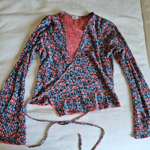 Blommig omlottopp från Pimkie - Säljer en blommig omlottopp från Pimkie med långa ärmar och knytband i midjan. Toppens mönster har röda, blå och svarta toner och materialet är mjukt och lätt. V-ringad och med plisserade detaljer framtill för extra stilpoäng.