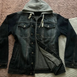 Svart jeansjacka med grå hoodie - Snygg svart jeansjacka med tvättad look och grå huva i sweatshirtmaterial. Jackan har knappar framtill, två bröstfickor med lock och snörning i luvan. Perfekt för dig som gillar streetstyle och vill ha en avslappnad vibe.