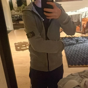 Grå hoodie från Stone Island Junior - Säljer en grå hoodie från Stone Island Junior med klassisk logotyp-patch på vänster ärm. Tröjan har huva, dragkedja framtill och ribbade muddar. Perfekt för dig som gillar streetwear och vill ha en clean look med snygga detaljer.
