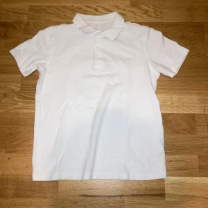 Vit pikétröja med krage och knappar - Klassisk vit pikétröja med korta ärmar, krage och tre vita knappar framtill. Tröjan är tillverkad i mjuk bomull som känns skön mot huden och har en stilren look som funkar till många olika outfits. Perfekt för dig som gillar enkel och clean stil.