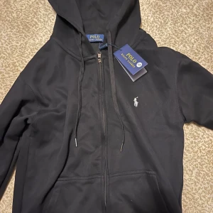 Svart hoodie från Polo Ralph Lauren - Svart hoodie från Polo Ralph Lauren med klassisk logga broderad på bröstet. Tröjan har huva med snörning, dragkedja framtill och två fickor. Materialet är mjuk bomull och passformen är normal. Perfekt för chill dagar och enkel att matcha med jeans.