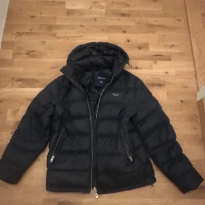 Svart pufferjacka från GANT med huva - Svart pufferjacka från GANT med huva och två sidofickor med dragkedja. Jackan har quiltad design och logga på bröstet. Insidan har en extra ficka med dragkedja och vita detaljer. Perfekt för kalla dagar och riktigt snygg stil.