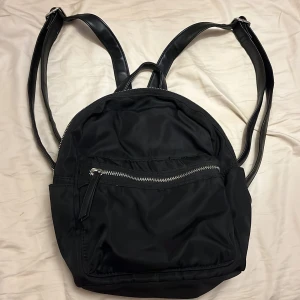 Svart ryggsäck med dragkedjor - En stilren svart ryggsäck med flera fack och silvriga dragkedjor. Väskan har justerbara axelremmar i syntetmaterial och ett rymligt huvudfack med innerfickor för extra förvaring. Perfekt för skola, jobb eller utflykter.