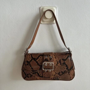 Brun axelväska med ormmönster H&M - Snygg axelväska från H&M i brunt ormmönstrat syntetmaterial. Väskan har ett brett axelband, silverfärgat spänne framtill och magnetlås. Insidan är fodrad i beige och har en innerficka. Perfekt för att lyfta din outfit. Helt ny med pris lapp! Nypris: 299kr