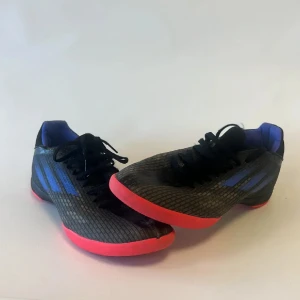 Svarta Adidas fotbollsskor med blå detaljer - Säljer ett par svarta Adidas fotbollsskor med blå ränder på sidorna och en snygg röd sula. Skorna har snörning och en låg profil, perfekt för inomhusspel. Insidan är fodrad med blått tyg för extra komfort. Grym stil för dig som gillar att sticka ut på planen.