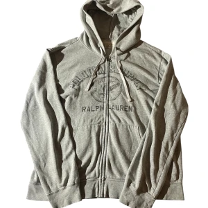Grå hoodie från Ralph Lauren Denim & Supply | XL - Snygg grå hoodie från Ralph Lauren Denim & Supply med tryck på bröstet. Tröjan har huva med snörning, dragkedja framtill och är fodrad med mjuk fleece. Perfekt för chill dagar och har en klassisk, loose passform.