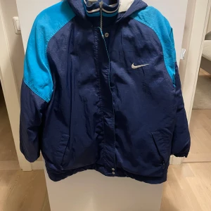 Blå och turkos täckjacka från Nike - Retrojacka från Nike i mörkblå och turkos färg med stor logga på ryggen och liten swoosh på bröstet. Jackan har huva, dragkedja och tryckknappar framtill samt två fickor. Perfekt för dig som gillar sportig streetstyle.