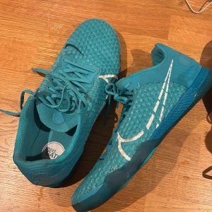 Nike React Gato turkosa futsalskor - Nike React Gato futsalskor i turkos med stickad ovandel och vita detaljer. Skorna har snörning, platt sula med cirkulära mönster och stor Nike-logga på sidan och undersidan. Perfekt för dig som gillar sportig stil och vill sticka ut med färg.