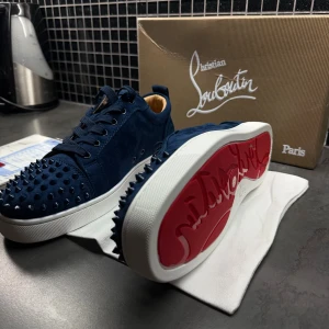 Mörkblå Christian Louboutin sneakers - Säljer ett par mörkblå sneakers från Christian Louboutin i mocka med ikoniska nitar på tån och sidorna. Skorna har snörning, vit platt sula och den klassiska röda undersidan. Insidan är beige och de levereras med originalkartong och dustbag.